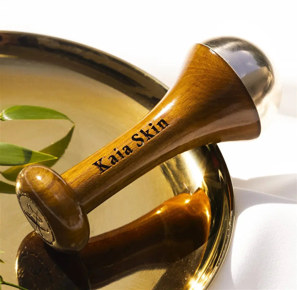 Kaia Skin Kansa Wand on a linen cloth beside face oil bottles, brass bowl, and Ayurvedic ritual setup